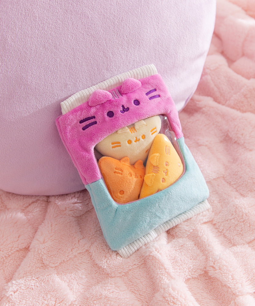 Pink and blue bag of plush sits on a fluffy surface. Pusheen the Cat's two eyes, mouth, and whiskers are embroidered in a dark purple thread above the see-through window in the middle front of the plush bag.