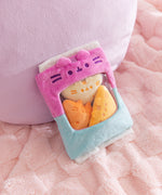 Pink and blue bag of plush sits on a fluffy surface. Pusheen the Cat's two eyes, mouth, and whiskers are embroidered in a dark purple thread above the see-through window in the middle front of the plush bag.