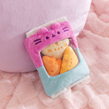 Pink and blue bag of plush sits on a fluffy surface. Pusheen the Cat's two eyes, mouth, and whiskers are embroidered in a dark purple thread above the see-through window in the middle front of the plush bag.