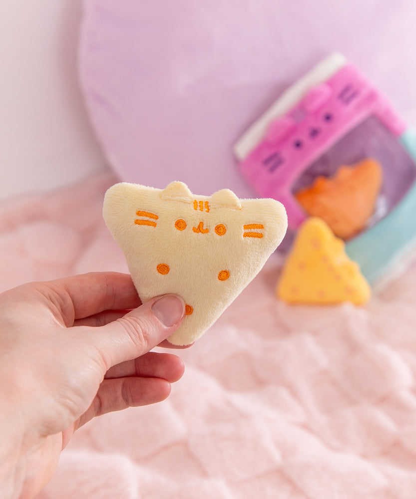 Gamer Pusheen Bag of Chips Plush sits on a white desktop. The pink and blue two-toned bag has a see-through window where the three yellow triangle plush reside.