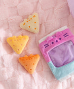 Three triangle plush in a range of yellow colors sit next the the plush bag they come in. Each chip plush has Pusheen's two ears extending off the plush and three stripes embroidered between the ears.