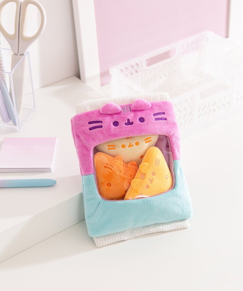 Pusheen Gamer Chips Plush its on a white desktop. The bag features white ribbed edged details on the top and bottom of the  two-toned blue and purple bag.