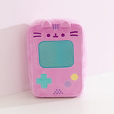 Front view of the pink rectangular shaped plush that mimics the look of a vintage gaming console. At the top of the plush toy, Pusheen's face is embroidered in dark purple while her three head stripes are embroidered in a medium purple color between her two plush ears. 