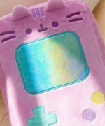 Gamer Pusheen Handheld Console Plush lies on a white surface. Pusheen the Cat takes the form of a pink retro gaming console with a large blue screen and buttons near the bottom of the plush.