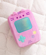 Gamer Pusheen Handheld Console Plush lies on a white surface. Pusheen the Cat takes the form of a pink retro gaming console with a large blue screen and buttons near the bottom of the plush.