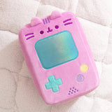 Gamer Pusheen Handheld Console Plush lies on a white surface. Pusheen the Cat takes the form of a pink retro gaming console with a large blue screen and buttons near the bottom of the plush.