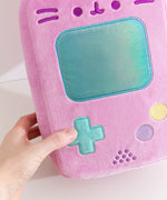 Model holds the edge of the Retro Handheld Console plush. The model's thumb touches the blue plus-sign button embroidered near the bottom left of the front of the rectangle shaped plush. 