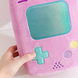Model holds the edge of the Retro Handheld Console plush. The model's thumb touches the blue plus-sign button embroidered near the bottom left of the front of the rectangle shaped plush. 