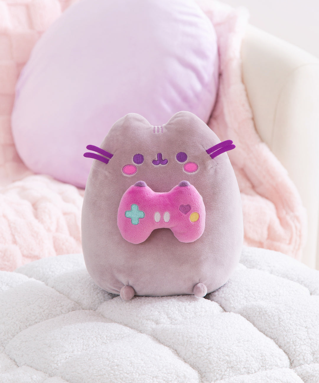 A pastel purple greay cat sits upright holding a pink gaming controller in her front paws. 