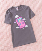 Pusheen t-shirt lies on a pink textured surface. The grey tee has a large graphic in the center front of the t-shirt featuring a pastel colored cat on the screen of a handheld gaming console. 