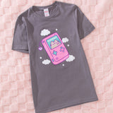 Pusheen t-shirt lies on a pink textured surface. The grey tee has a large graphic in the center front of the t-shirt featuring a pastel colored cat on the screen of a handheld gaming console. 