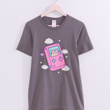 Gamer Pusheen Retro Handheld Tee hangs on a light pink hanger on a light background. The grey tee shirt has a graphic of Pusheen the Cat inside a pink handheld gaming console surrounded by fluffy white clouds and stars. 