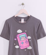 Dark gray t-shirt with a large graphic on the front center. The graphic shows Pusheen the Cat inside a pink gaming device with blue and purple plus sign and circle shaped buttons. 