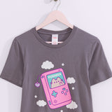 Dark gray t-shirt with a large graphic on the front center. The graphic shows Pusheen the Cat inside a pink gaming device with blue and purple plus sign and circle shaped buttons. 
