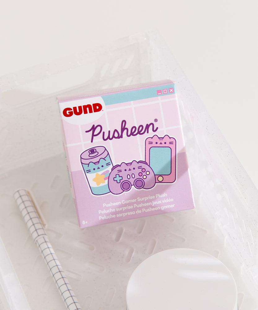 A close-up of the Gamer Pusheen Surprise Plush packaging. The front of the packaging features Pusheen as various gaming characters including a striped energy drink, purple controller, and pink handheld gaming console. The packaging says "Pusheen Gamer Surprise Plush."