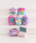Seven gaming themed mini plush keychains sit atop a white pedestal. The assortment includes Pusheen the Cat taking the form of an accessory case, a bag of chips, two different handheld gaming consoles, and energy drink, a controller, and a laptop.