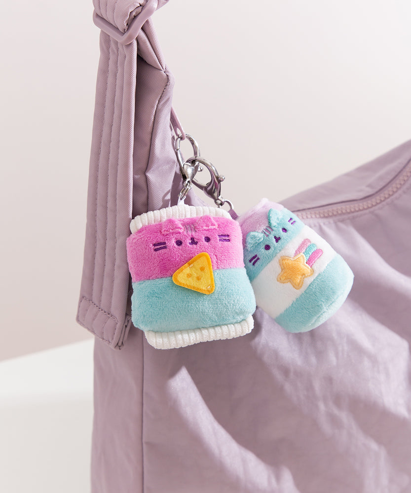Close-up shot of Gamer Pusheen accessories: a bag of chips and an energy drink. The two blue and pink keychains are attached to a zipper on a purple handbag.  