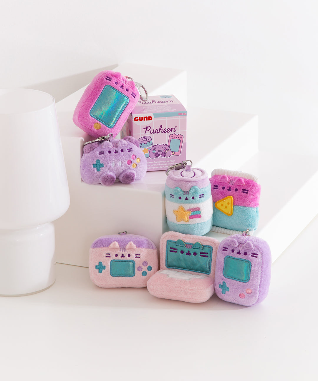 Front view of Gamer Pusheen Surprise Plush assortment. Plush keychains sit on multi-level white pedestal in front of a white background. Keychains feature various gaming accessories including handheld gaming consoles, controller, energy drink, and laptops. Accompanying the seven gamer keychains is the mystery box packaging for the Gamer Surprise Plush.