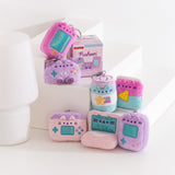 Front view of Gamer Pusheen Surprise Plush assortment. Plush keychains sit on multi-level white pedestal in front of a white background. Keychains feature various gaming accessories including handheld gaming consoles, controller, energy drink, and laptops. Accompanying the seven gamer keychains is the mystery box packaging for the Gamer Surprise Plush.