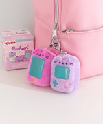 Close-up shot of Pusheen Gaming consoles. The plush keychains are attached to a pink bag. Next to the purple and pink plushies are the Gamer surprise plush packaging box. 