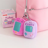 Close-up shot of Pusheen Gaming consoles. The plush keychains are attached to a pink bag. Next to the purple and pink plushies are the Gamer surprise plush packaging box. 