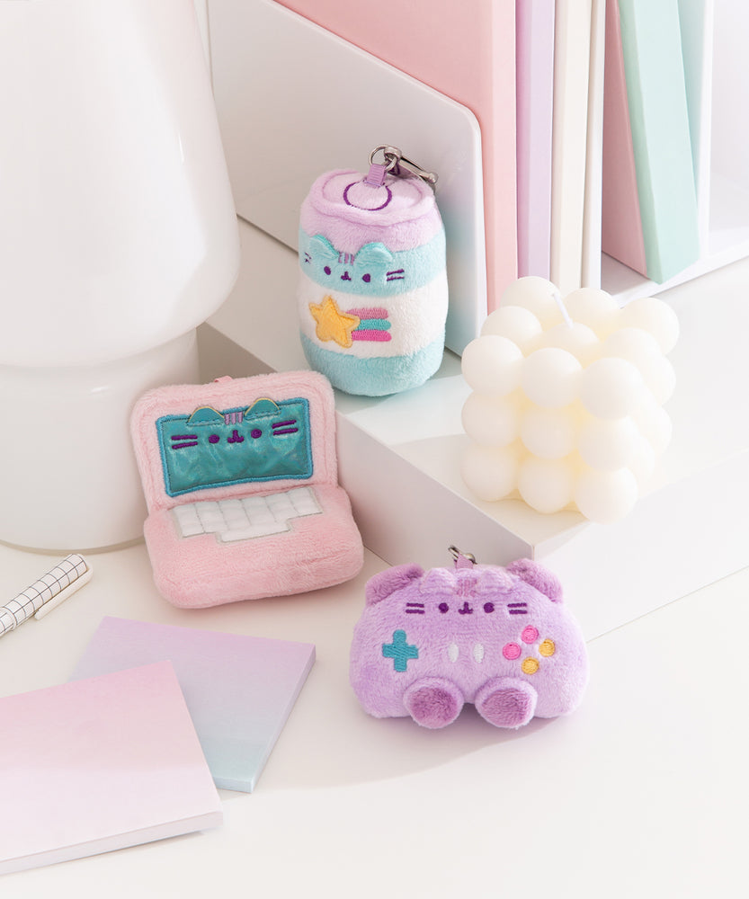 Energy drink, laptop, and controller mini keychains sits on a desktop. The collection of miniature plush have a light pink, purple, and blue color palette.  