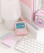 Mini laptop plush sits atop a white desktop with various accessories surrounding the keychain. The light pink laptop has a white keyboard and an iridescent teal screen. 