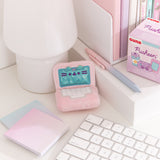 Mini laptop plush sits atop a white desktop with various accessories surrounding the keychain. The light pink laptop has a white keyboard and an iridescent teal screen. 
