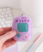 Model holds handheld gaming console keychain with Pusheen’s facial features. The light purple rectangle has an iridescent teal square in the middle of the miniature plush keychain.  