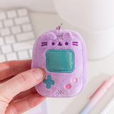 Model holds handheld gaming console keychain with Pusheen’s facial features. The light purple rectangle has an iridescent teal square in the middle of the miniature plush keychain.  