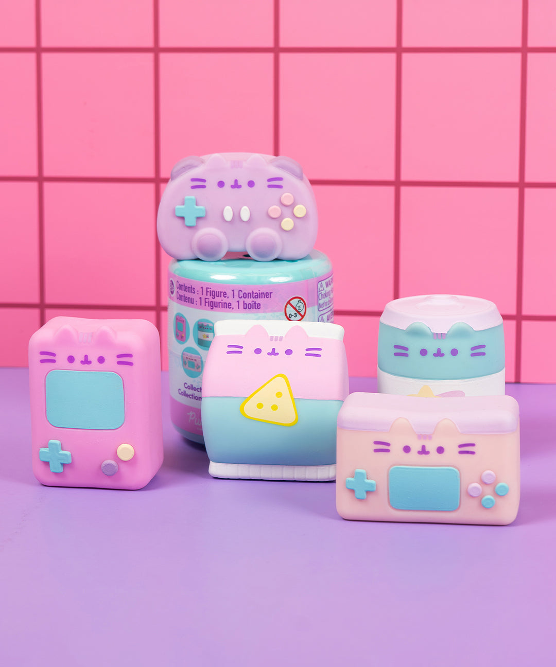 Front view of all Gamer Pusheen Surprise Squishy including the blind box capsule. The squishies include a retro handheld console, a gaming controller, and bag of chips, a soda can, and a modern handheld gaming console.