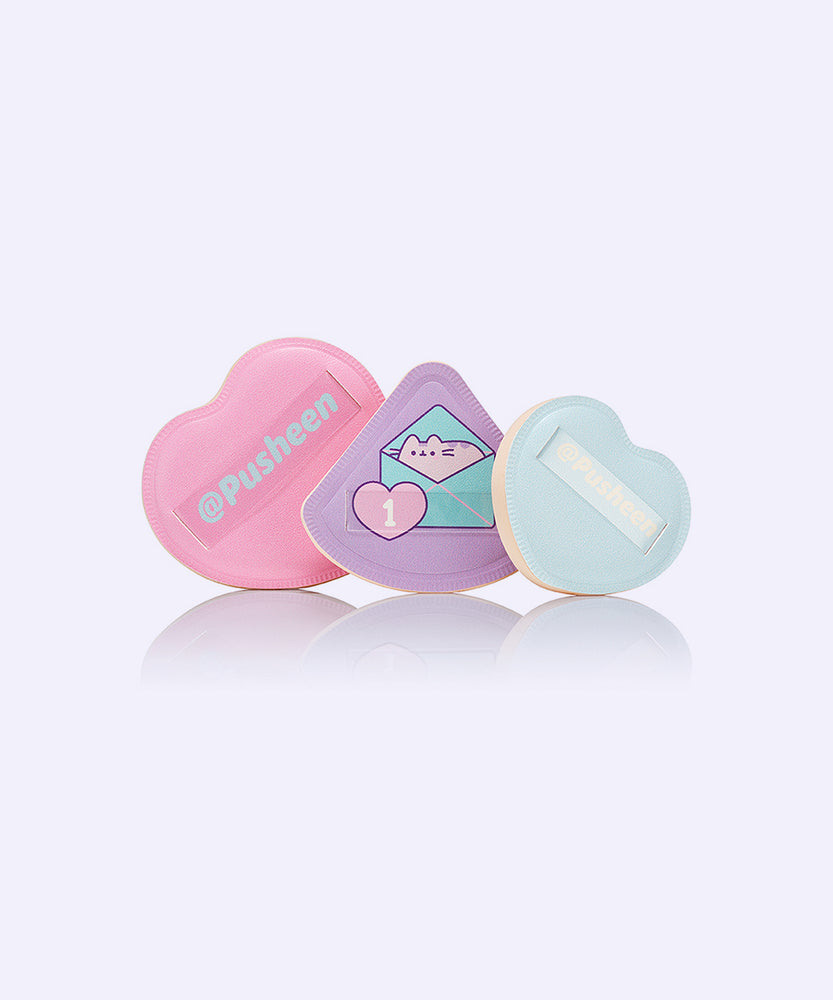 Puff trio includes three different colors and sizes. The pink puff is in the shape of a heart. The light purple puff is a triangle with a graphic of Pusheen in an envelope on the holding side. The light blue puff is a smaller heart shape than the pink puff.  