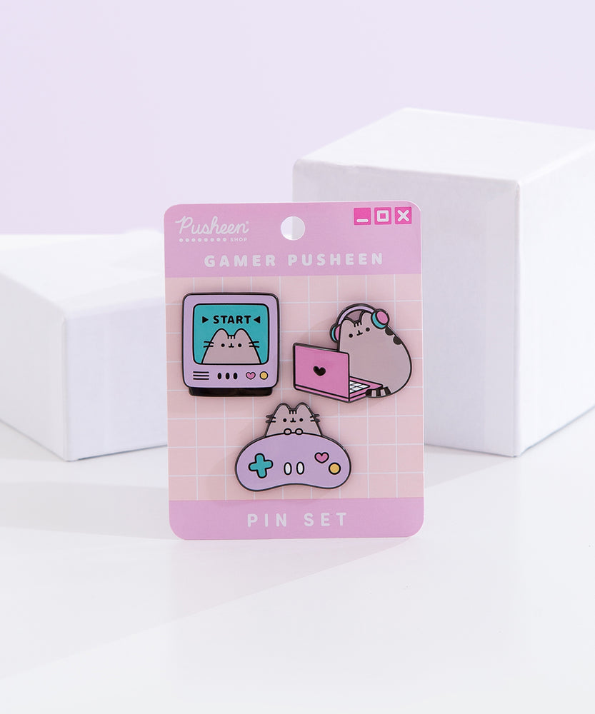 Gamer Pusheen Pin Set lies in front of two white blocks and a light purple background. The three Pusheen pins are attached to a backer card with the product name in a white color. The top left pin shows Pusheen in a gaming monitor. The top right pin shows the cat with headphones on in front of a laptop.  The bottom pin shows Pusheen with a light purple gaming controller.  