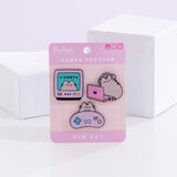 Gamer Pusheen Pin Set lies in front of two white blocks and a light purple background. The three Pusheen pins are attached to a backer card with the product name in a white color. The top left pin shows Pusheen in a gaming monitor. The top right pin shows the cat with headphones on in front of a laptop.  The bottom pin shows Pusheen with a light purple gaming controller.  