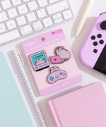 A close-up view of the three Pusheen pins on their backer card lying on a white desk surrounded by desk accessories. Surrounding the pin set is a gaming controller, two notebooks, two pens, and a keyboard.   