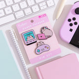 A close-up view of the three Pusheen pins on their backer card lying on a white desk surrounded by desk accessories. Surrounding the pin set is a gaming controller, two notebooks, two pens, and a keyboard.   