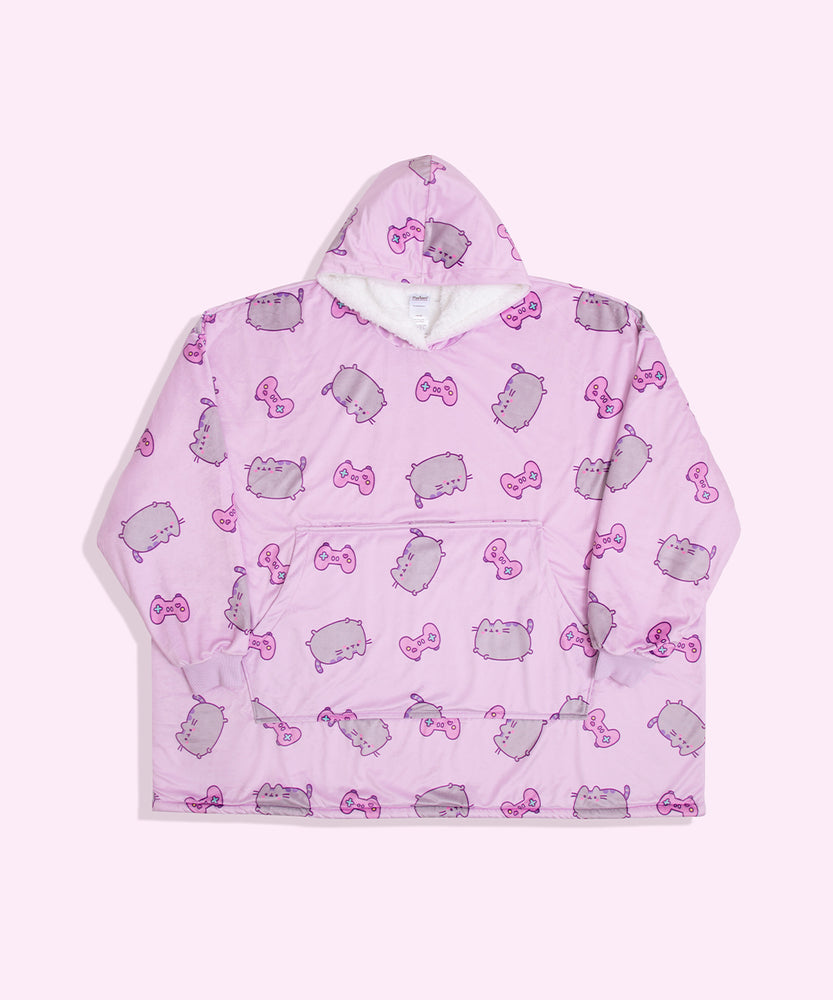 Gamer Pusheen Wearable Blanket Hoodie lies on a light surface. The oversized pullover has a hood, oversized sleeves, a large front pocket, and an all-over-print Pusheen the Cat with pink gaming controllers.