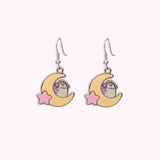 Front view of the Gaming Pusheen Dangle Earrings. Wearing pink and mint green headphones, Pusheen the Cat has light purple back stripes while sitting on a yellow crescent moon with a light pink star.  