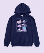 A navy-blue graphic hoodie lies against a light purple background. The unisex hoodie has a screen-printed graphic on the center of the chest showing Pusheen the Cat outlined in purple with her gaming session essentials printed in pastel purples, pinks, and blues. 