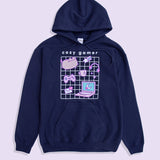 A navy-blue graphic hoodie lies against a light purple background. The unisex hoodie has a screen-printed graphic on the center of the chest showing Pusheen the Cat outlined in purple with her gaming session essentials printed in pastel purples, pinks, and blues. 