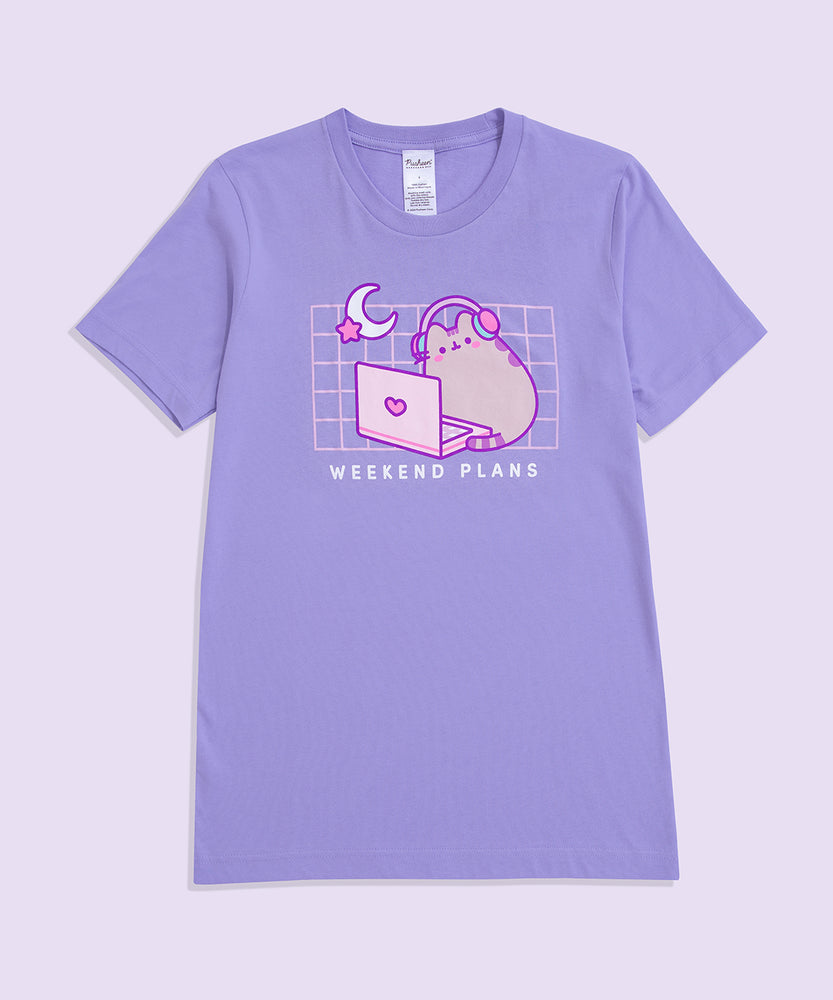 A purple graphic t-shirt lies against a light purple background. The unisex tee shirt has a screen-printed graphic in the center of the chest showing Pusheen the Cat with headphones on sitting in front of a laptop. 