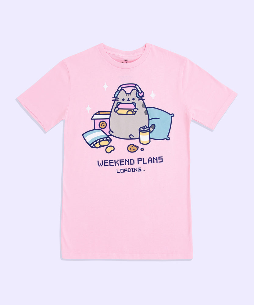 A light pink graphic tee against a light purple background. The center graphic features Pusheen wearing headphones over her ears and holding a pink, yellow, and blue gaming controller surrounded by chips, cookies, and pillows. Under Pusheen is the text “Weekend Plans Loading...”  