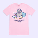 A light pink graphic tee against a light purple background. The center graphic features Pusheen wearing headphones over her ears and holding a pink, yellow, and blue gaming controller surrounded by chips, cookies, and pillows. Under Pusheen is the text “Weekend Plans Loading...”  