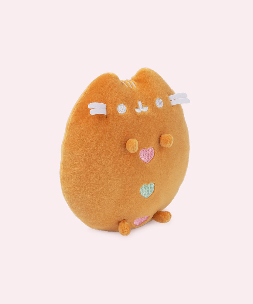 Gingerbread Squisheen Plush sits in front of a white background and gold present bows. The light brown plush has four paws that extend off the body. The cat’s white facial features are embroidered in white thread.  