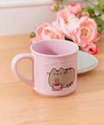 Pink mug with a Pusheen design and 'good morning' text on a wooden table with flowers in the background.