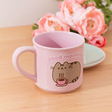 Pink mug with a Pusheen design and 'good morning' text on a wooden table with flowers in the background.