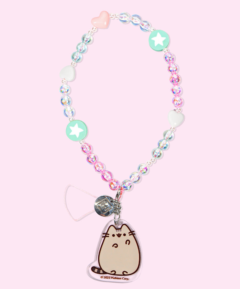The Pusheen Beaded Phone Charm lies on a pink background. The beaded string includes pink and clear round beads, mint green round beads with white stars, and white and light pink heart-shaped beads. At the end of the string is an acrylic charm of Pusheen the Cat waving.