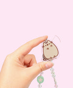 Model holds Pusheen charm at the end of the beaded phone charm. Pusheen the Cat is shown in grey with dark brown features and outline. The figure is set on a clear background.  
