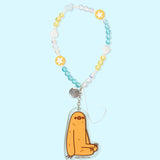    The Sloth Beaded Phone Charm lies on a blue background. The beaded string includes blue, yellow, and clear round beads, yellow round beads with white stars, and white and light blue heart-shaped beads. At the end of the string is an acrylic charm of Pusheen’s friend Sloth sitting.  