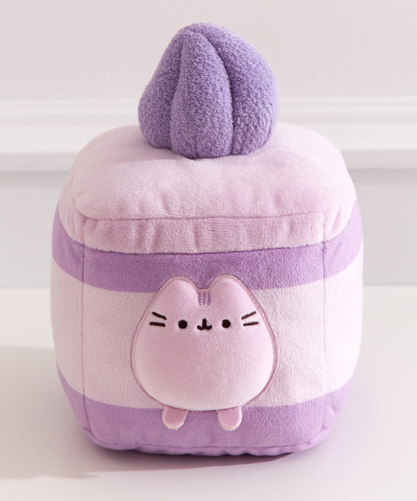 Purple cake plush lies on a pink textured surface. Pusheen transforms into a layered lavender-flavored mini cake. 